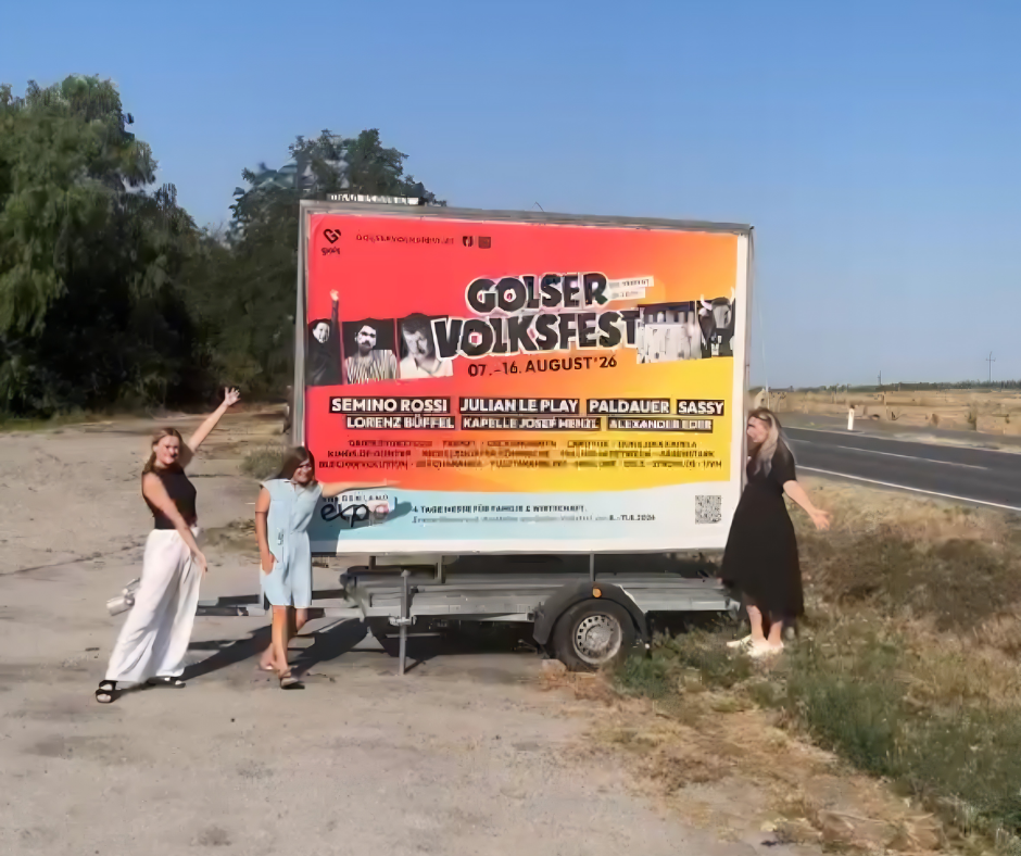 Ein sonnenbeschienenes Foto zeigt drei Frauen, die neben einem großen, auf einem Anhänger montierten Plakat für das "Golser Volksfest" posieren.